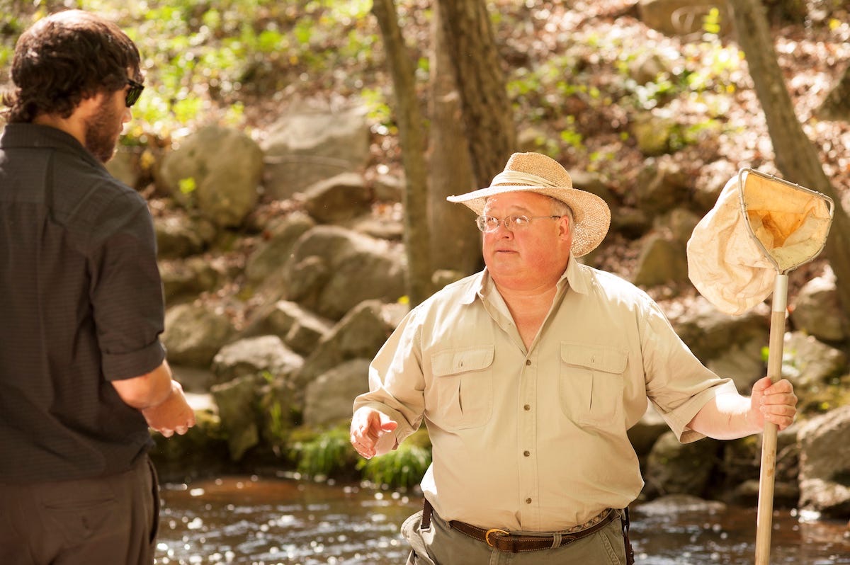 Dr. Groover standing in a shallow forest stream holding a dip net, talking with a younger man
