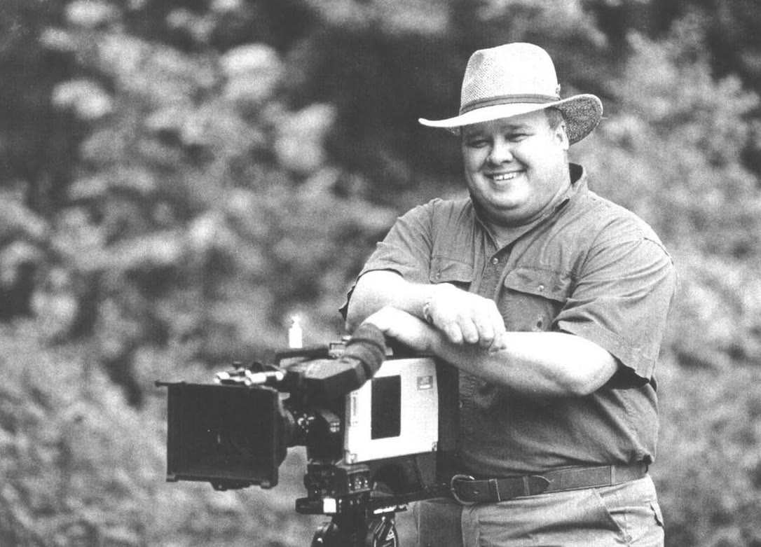 Richard Groover in a wide-brimmed hat, smiling and leaning on a professional video camera outdoors