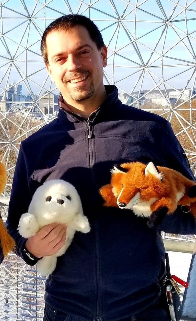 Norm Leo smiling inside the geodesic dome of the Montréal Biosphere, holding plush toys of a harp seal and a fox