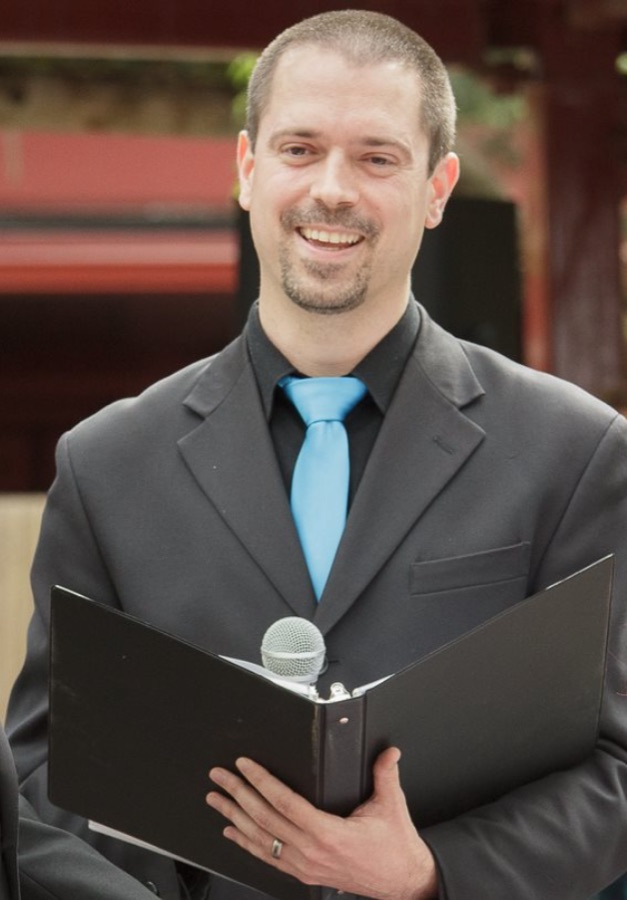 Norm Leo smiling at a microphone, holding a binder of notes