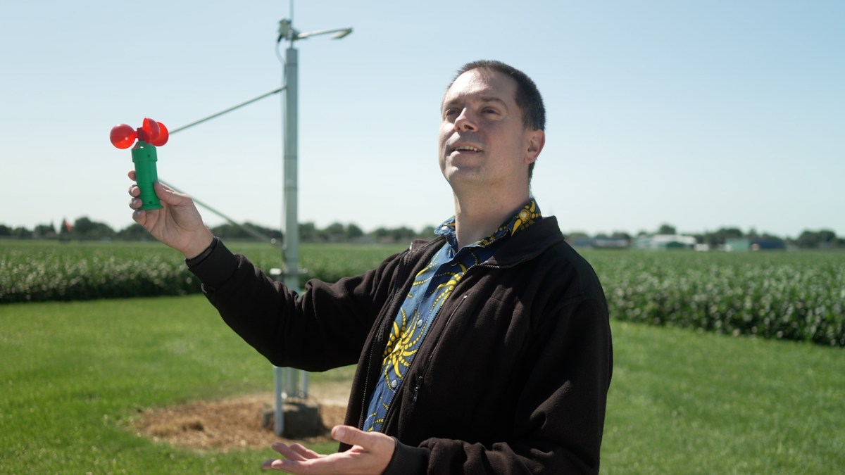 Norm Leo outdoors in a green field next to a weather station, holding up a small anemometer